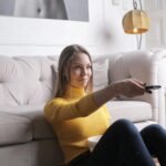Adult woman in yellow sweater lounging on plush sofa holding TV remote, enjoying leisure time indoors.