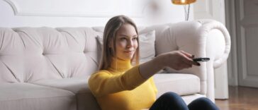 Adult woman in yellow sweater lounging on plush sofa holding TV remote, enjoying leisure time indoors.