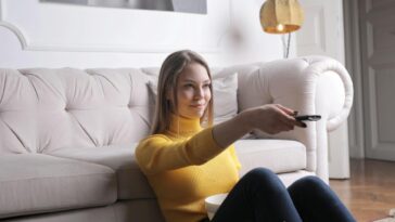Adult woman in yellow sweater lounging on plush sofa holding TV remote, enjoying leisure time indoors.