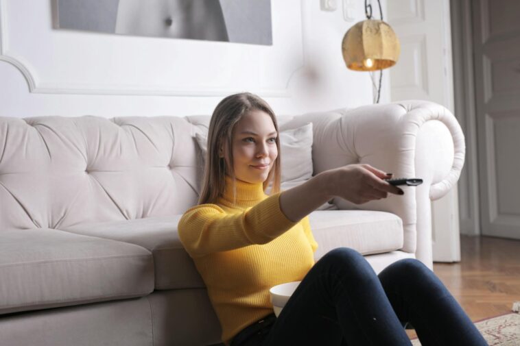 Adult woman in yellow sweater lounging on plush sofa holding TV remote, enjoying leisure time indoors.