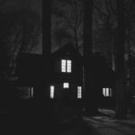 A haunting black and white image of a house surrounded by trees at night, showcasing an eerie atmosphere.