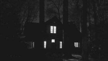 A haunting black and white image of a house surrounded by trees at night, showcasing an eerie atmosphere.