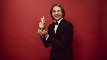 Smiling man in a tuxedo holding a trophy against a red background, celebrating success.