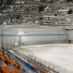 Wide-angle view of an empty indoor ice rink with visible steel beams and seating area.