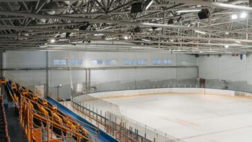 Wide-angle view of an empty indoor ice rink with visible steel beams and seating area.