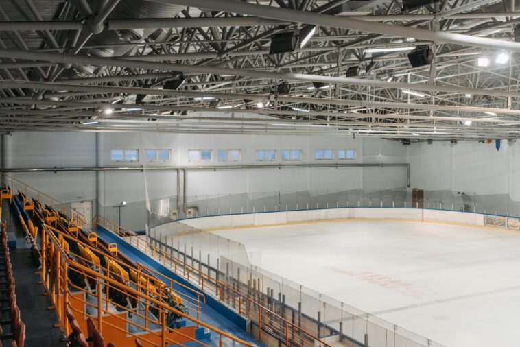 Wide-angle view of an empty indoor ice rink with visible steel beams and seating area.