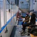 Father and son sitting on a bench at an ice skating rink, watching a game in progress.
