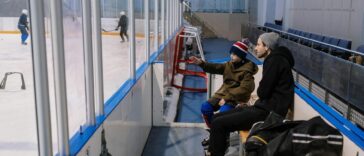 Father and son sitting on a bench at an ice skating rink, watching a game in progress.