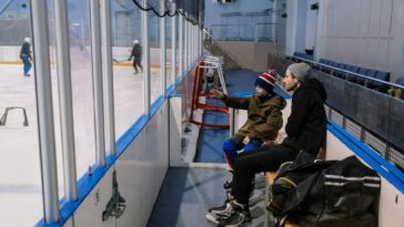 Father and son sitting on a bench at an ice skating rink, watching a game in progress.