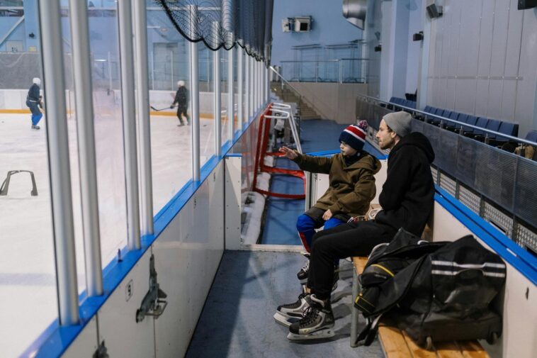 Father and son sitting on a bench at an ice skating rink, watching a game in progress.
