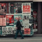 Side view of senior man in casual clothes walking along wall of modern building with posters while having stroll on city street