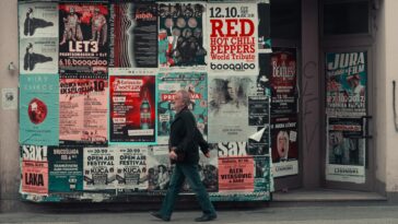 Side view of senior man in casual clothes walking along wall of modern building with posters while having stroll on city street