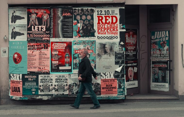 Side view of senior man in casual clothes walking along wall of modern building with posters while having stroll on city street