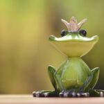 A cute frog prince figurine sits gracefully outdoors with a blurred green background.