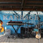 An outdoor stage with various musical instruments including a cello, drum set, and keyboard.