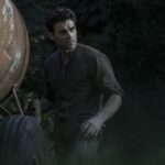 A man with short hair stands beside a rustic trailer in a forest setting.