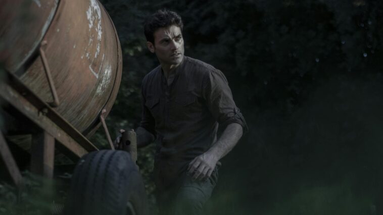 A man with short hair stands beside a rustic trailer in a forest setting.