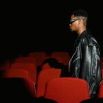 A man wearing glasses walks through a dimly lit movie theater with red seats.