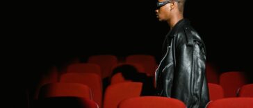 A man wearing glasses walks through a dimly lit movie theater with red seats.
