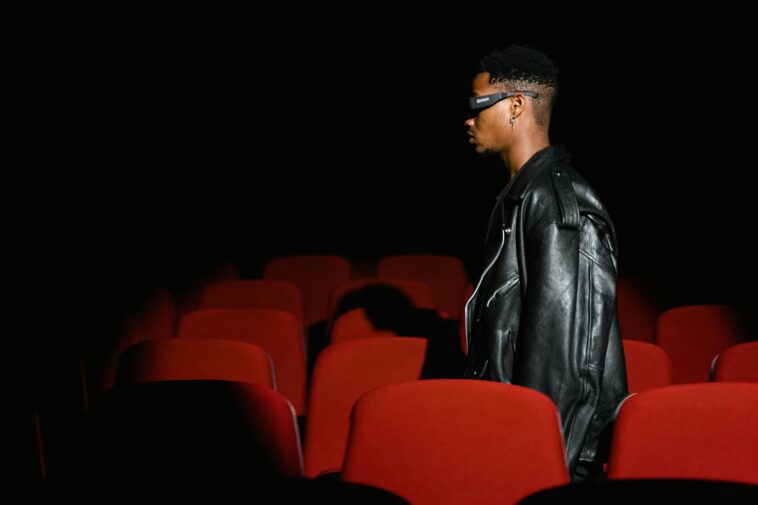 A man wearing glasses walks through a dimly lit movie theater with red seats.