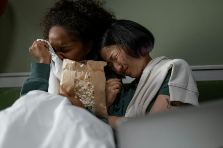 Two women sharing an emotional moment while watching a movie at home, enjoying popcorn.