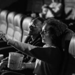 A couple laughing and enjoying a movie in a dimly lit theater, sharing popcorn.