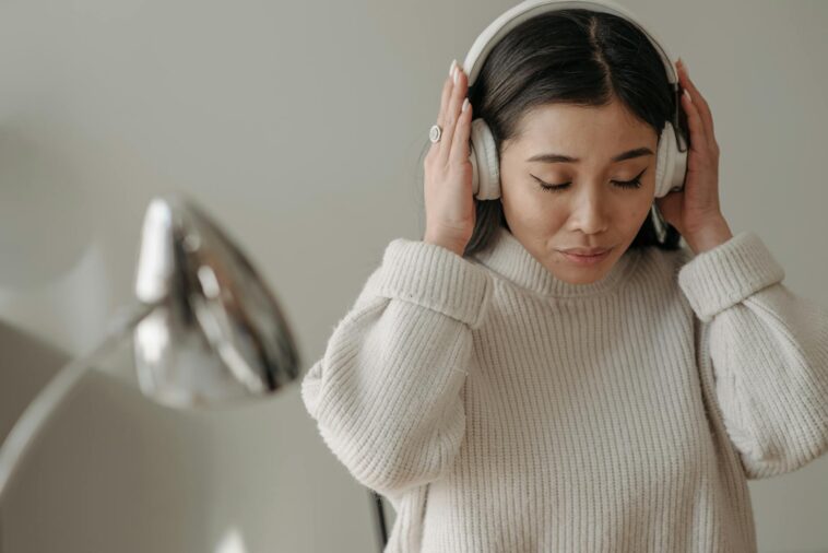 Asian woman wearing headphones and a knitted sweater, enjoying music indoors.