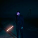 Dark night scene of a mysterious figure in a glowing mask holding a light sword.