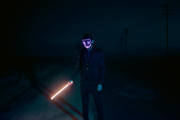 Dark night scene of a mysterious figure in a glowing mask holding a light sword.