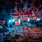 Side view of ethnic vendor and unrecognizable client standing near street stall with assorted meat barbecue on skewers under glowing lamps in evening