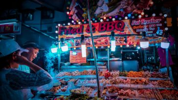 Side view of ethnic vendor and unrecognizable client standing near street stall with assorted meat barbecue on skewers under glowing lamps in evening