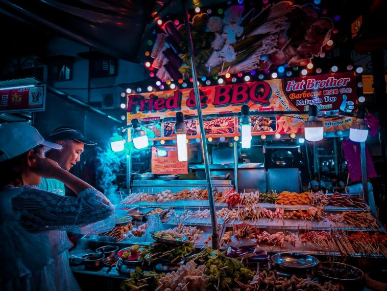 Side view of ethnic vendor and unrecognizable client standing near street stall with assorted meat barbecue on skewers under glowing lamps in evening