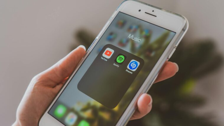 Hand holding a smartphone displaying popular music apps like Spotify and Shazam in a close-up shot.