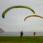 Two paragliders on a grassy cliff by the ocean, ready for flight adventure.