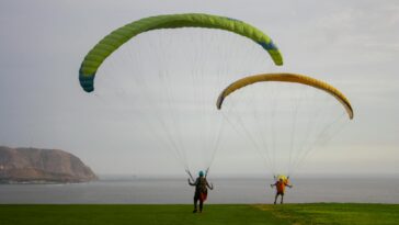 Two paragliders on a grassy cliff by the ocean, ready for flight adventure.
