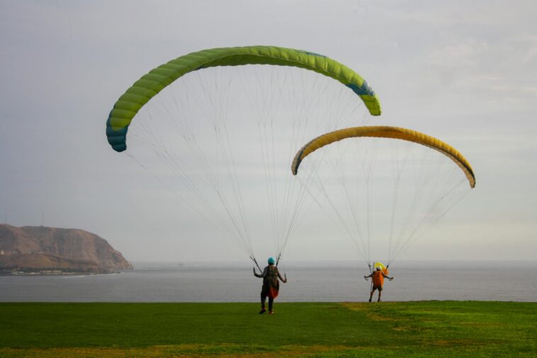 Two paragliders on a grassy cliff by the ocean, ready for flight adventure.