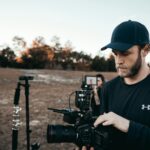 A bearded man adjusting camera equipment outdoors with a film crew in the background.