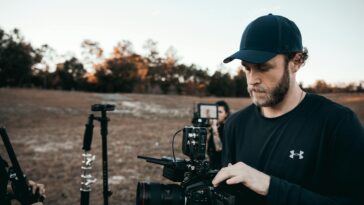 A bearded man adjusting camera equipment outdoors with a film crew in the background.