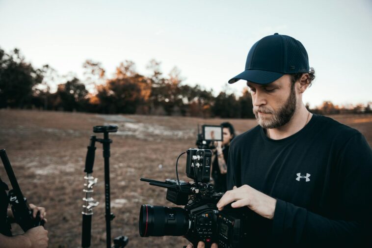 A bearded man adjusting camera equipment outdoors with a film crew in the background.