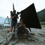 Behind-the-scenes view of film production with actors and crew on location outdoors.