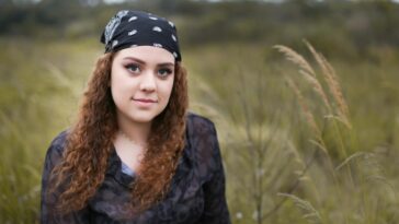 A young woman with curly hair and a bandana poses outdoors in a grassy field.
