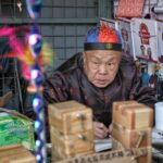Elderly man in traditional attire working at a market stall in Beijing, China.