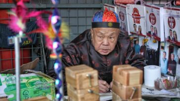 Elderly man in traditional attire working at a market stall in Beijing, China.