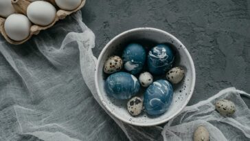 A collection of artistically dyed Easter eggs in a bowl, set on a textured concrete surface with rustic decor.