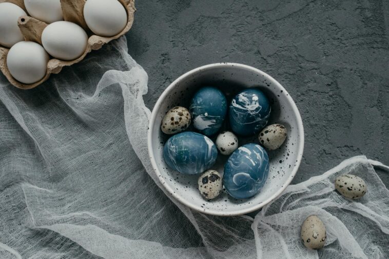 A collection of artistically dyed Easter eggs in a bowl, set on a textured concrete surface with rustic decor.