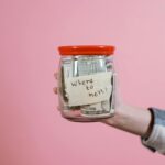 A hand holding a glass jar filled with dollar bills labeled 'Where to next?' against a pink background, symbolizing savings and future planning.