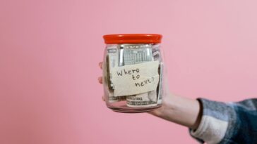 A hand holding a glass jar filled with dollar bills labeled 'Where to next?' against a pink background, symbolizing savings and future planning.