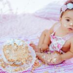 Cute baby enjoying a cake smash for her first birthday, sitting outdoors on a blanket.