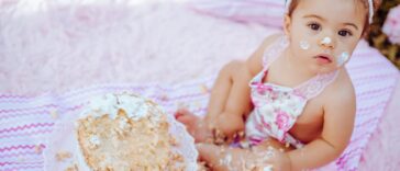 Cute baby enjoying a cake smash for her first birthday, sitting outdoors on a blanket.