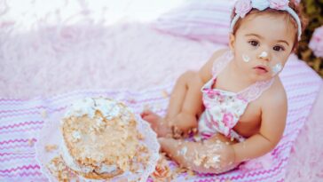 Cute baby enjoying a cake smash for her first birthday, sitting outdoors on a blanket.